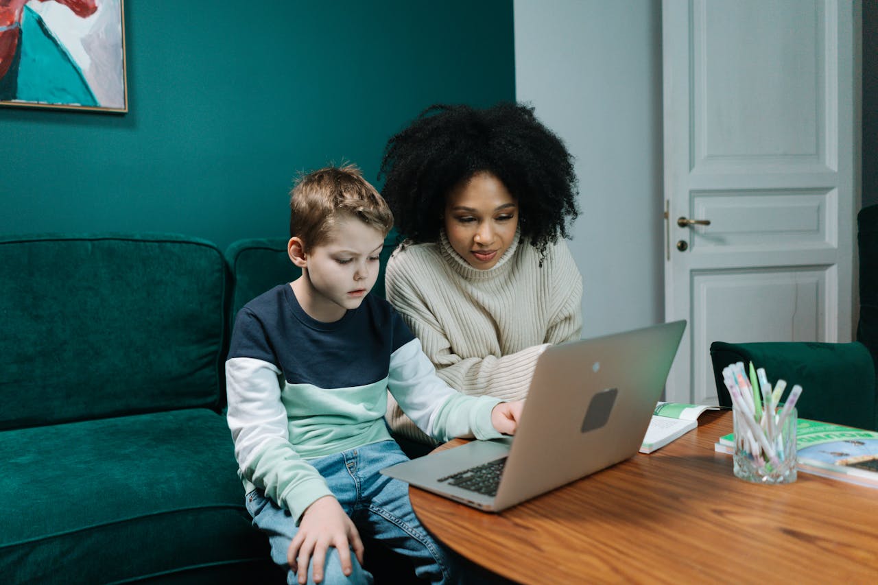 A woman and a child sit together on a sofa as they work on homework using a laptop.